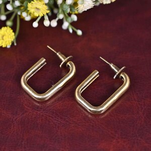 Könnte beinhalten: Ein Paar goldfarbene Creolen mit quadratischer Form. Die Ohrringe haben ein schlichtes, minimalistisches Design.