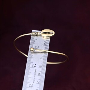 May include: A gold-toned metal cuff bracelet featuring a cowrie shell charm at one end and a small spherical bead at the other. A metal ruler is positioned across the bracelet, showing the shell charm measures approximately 1 inch in length.