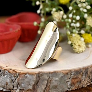 May include: A gold-toned, oval-shaped ring with a smooth, slightly textured surface. The ring is open on one side and has a band on the other side.
