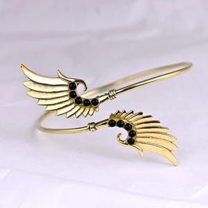 May include: A gold-toned bangle bracelet with two wing-shaped charms. Each wing is adorned with black beads.