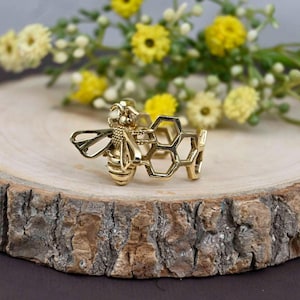 Puede incluir: Anillo dorado con un diseño detallado de abeja y patrón de panal. El anillo se encuentra sobre una superficie de madera, con flores amarillas en el fondo. Una joya con temática de la naturaleza, ideal para regalo.