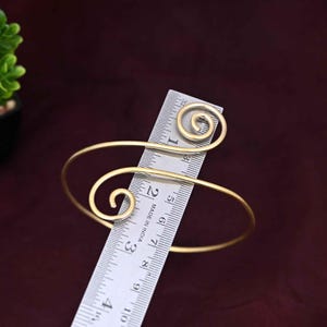 May include: A gold-toned metal arm cuff featuring two spiral ends, positioned against a metal ruler that shows a diameter of approximately 3 inches. The open-ended bracelet has a smooth, matte finish and a curved design that wraps around the arm.