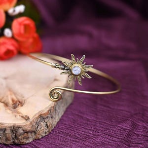 May include: A gold-colored cuff bracelet featuring a sunburst design with a central, light-colored stone. The bracelet has a spiral detail at one end and is displayed on a wooden surface against a purple background.