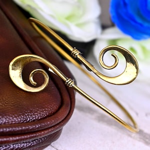 May include: A gold-colored arm cuff bracelet with a spiral design at each end. The bracelet has a simple, elegant design, with a thin band and decorative swirls. The cuff is shown against a brown leather surface and a blurred floral background.