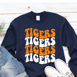May include: A navy blue sweatshirt with the word "TIGERS" repeated four times in a stacked design. The word "TIGERS" is in a retro font with orange and white letters. The sweatshirt is laying on a white wooden surface with a pair of white sneakers and blue jeans.