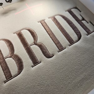 May include: Embroidered "BRIDE" in brown thread on a white fabric background.