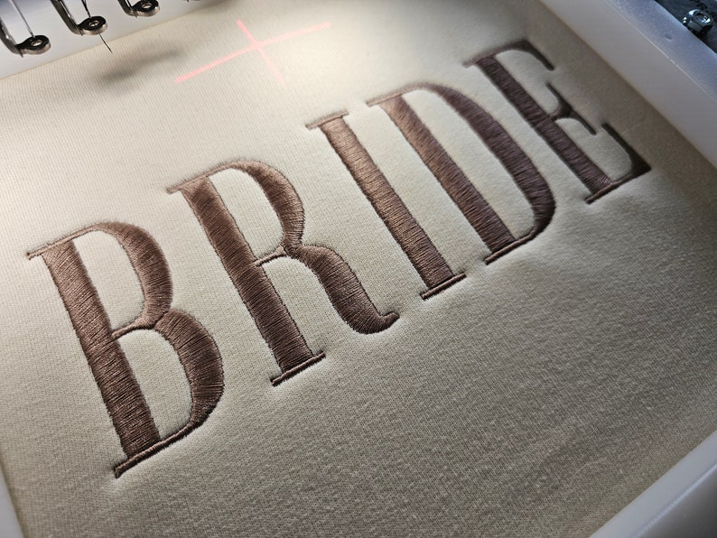May include: Embroidered "BRIDE" in brown thread on a white fabric background.