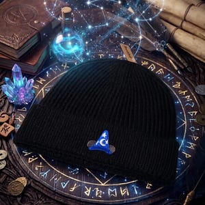May include: A black ribbed knit beanie featuring a small embroidered patch on the front. The patch depicts a blue wizard hat with white stars and a crescent moon, positioned between two small dark circular shapes resembling mouse ears.
