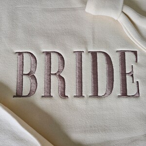 May include: A white sweatshirt with the word "BRIDE" embroidered in brown on the front.