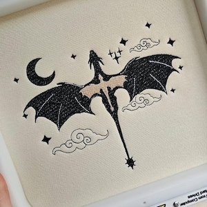 May include: Black embroidered design of a dragon flying in the night sky with a crescent moon, stars, and clouds. The design is on a cream-coloured fabric.
