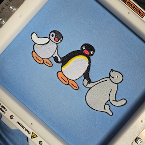 Puede incluir: Diseño bordado de tres animales de dibujos animados sobre una tela azul claro. El diseño presenta un pingüino con una franja amarilla en el pecho, un pingüino más pequeño con un pico rojo y una foca gris. Los animales se están dando la mano.