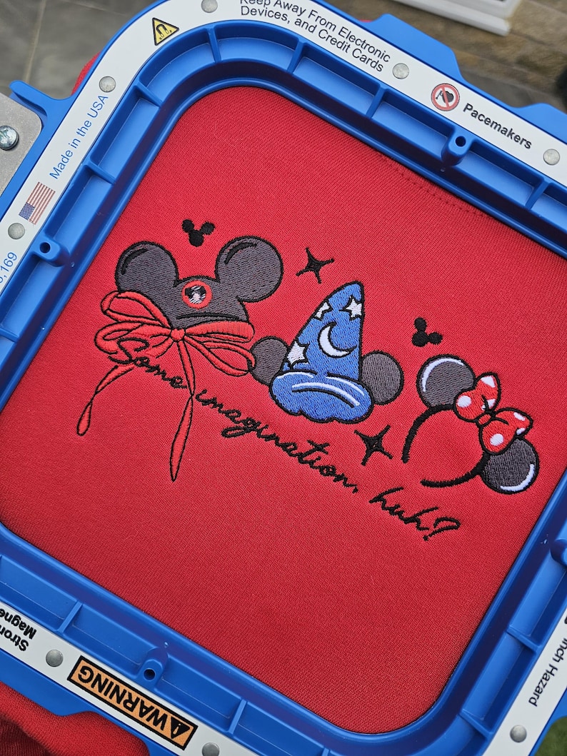 May include: Red fabric with embroidered cartoon character designs, including a mouse head with a bow, a wizard hat, and mouse ears with a bow. The text "Some imagination, huh?" is also embroidered.