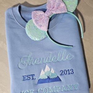 May include: A light blue sweatshirt with the text "Arendelle EST. 2013 ICE COMPANY" embroidered on the front. The sweatshirt is decorated with a light blue and pink sequined headband with a bow.
