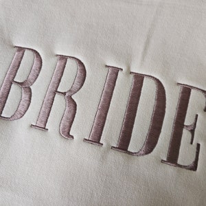 May include: A white fabric with the word "BRIDE" embroidered in a light brown color.