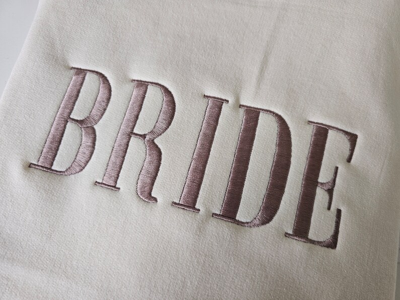 May include: A white fabric with the word "BRIDE" embroidered in a light brown color.