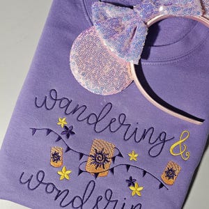 Peut inclure: Un sweat-shirt lavande avec la broderie "wandering & wondering" et un motif de lanternes et d'étoiles. Un serre-tête à oreilles de Minnie Mouse à paillettes roses et lavande est posé sur le sweat-shirt.
