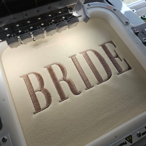 May include: A close-up of an embroidery machine with a beige fabric hoop. The word "BRIDE" is embroidered in a brown thread on the fabric.