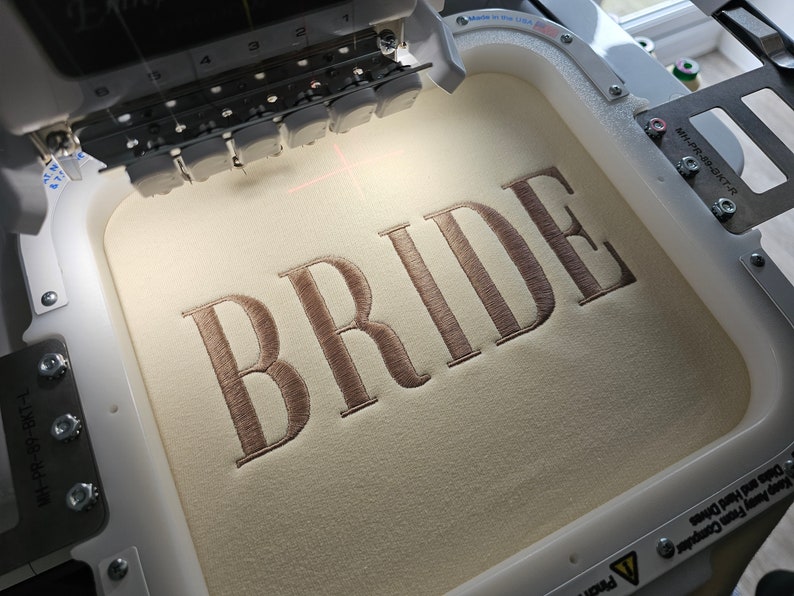 May include: A close-up of an embroidery machine with a beige fabric hoop. The word "BRIDE" is embroidered in a brown thread on the fabric.