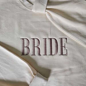 May include: A white sweatshirt with the word "BRIDE" embroidered in brown on the front.