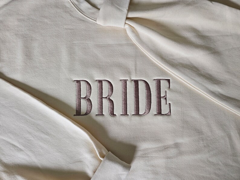 May include: A white sweatshirt with the word "BRIDE" embroidered in brown on the front.