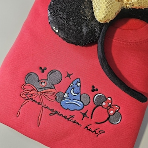 May include: A red sweatshirt with embroidered Mickey Mouse-themed designs and the text "Some imagination, huh?". A black sequined ear and a gold sequined bow headband are on top of the sweatshirt.