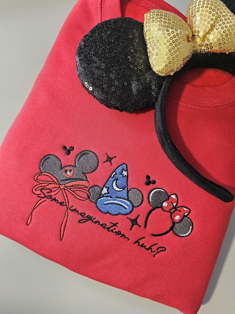 May include: A red sweatshirt with embroidered Mickey Mouse-themed designs and the text "Some imagination, huh?". A black sequined ear and a gold sequined bow headband are on top of the sweatshirt.