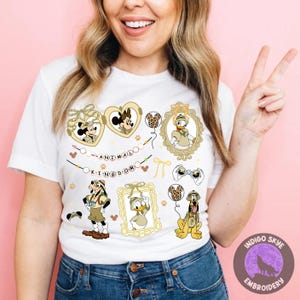Può includere: T-shirt bianca con i personaggi dei cartoni animati Topolino e Minnie, Paperino e Pluto, con la scritta "ANIMAL KINGDOM". Il design include cornici a forma di cuore, binocoli e impronte di zampe a tema safari.
