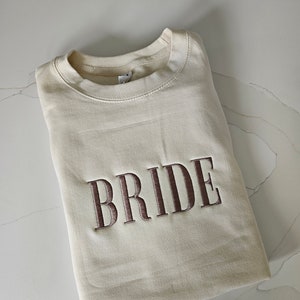 May include: A cream-colored sweatshirt with the word "BRIDE" embroidered in brown on the front.