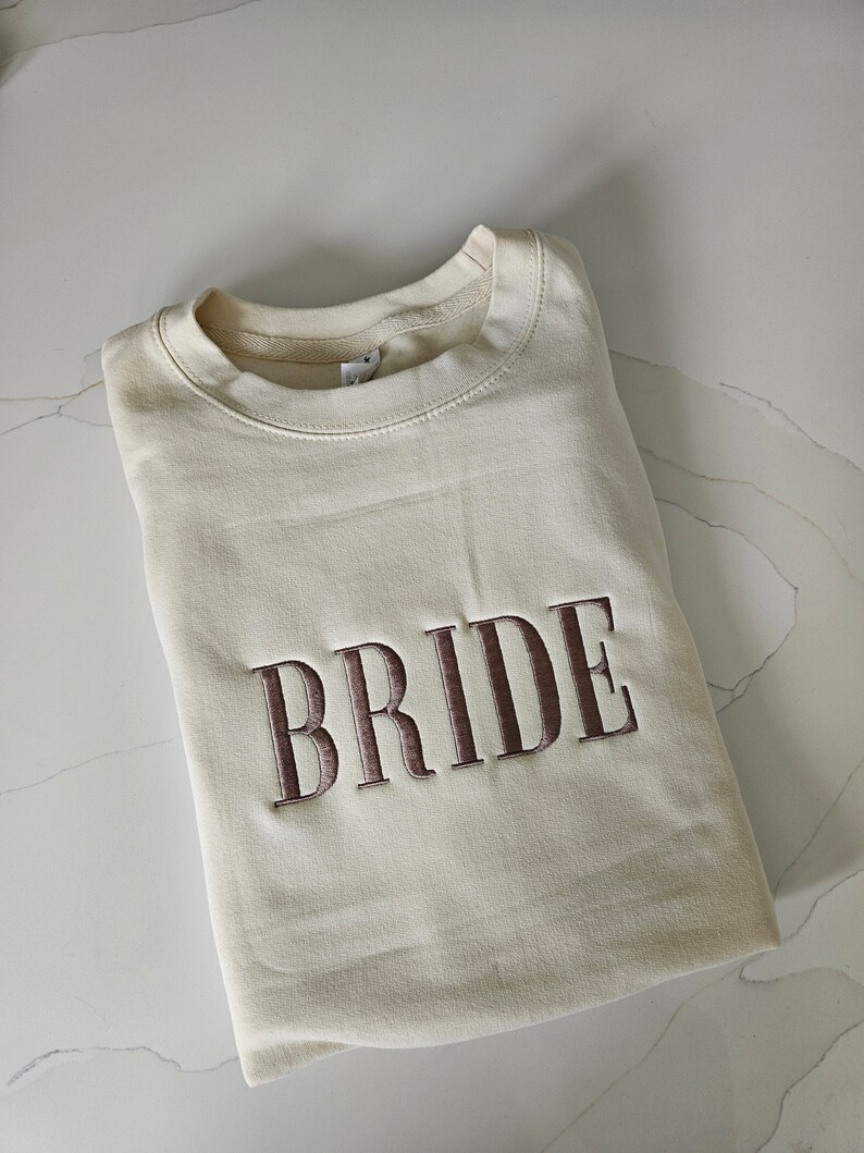 May include: A cream-colored sweatshirt with the word "BRIDE" embroidered in brown on the front.