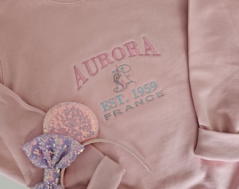 Könnte beinhalten: Rosafarbener Pullover mit dem gestickten Text "AURORA EST. 1959 FRANCE" auf der Vorderseite. Ein rosa paillettenbesetztes Stirnband mit Mäuseohren liegt auf dem Pullover.