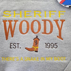 May include: Gray sweatshirt with embroidered text that reads "Sheriff Woody Est. 1995 There's a snake in my boot". The text is in yellow, orange, and brown thread.