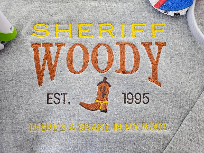 May include: Gray sweatshirt with embroidered text that reads "Sheriff Woody Est. 1995 There's a snake in my boot". The text is in yellow, orange, and brown thread.
