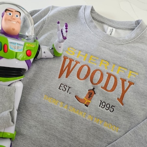May include: A gray sweatshirt with the text "Sheriff Woody Est. 1995 There's a snake in my boot" in orange and yellow embroidery. A toy Buzz Lightyear action figure is in the background.