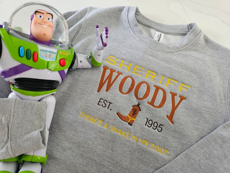 May include: A gray sweatshirt with the text "Sheriff Woody Est. 1995 There's a snake in my boot" in orange and yellow embroidery. A toy Buzz Lightyear action figure is in the background.