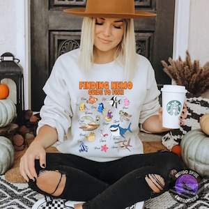 Peut inclure: Sweat-shirt blanc avec l'inscription "FINDING NEMO GUIDE TO FISH" en orange et noir. Le sweat-shirt présente des illustrations de personnages de poissons du film. La personne tient une tasse à café blanche.
