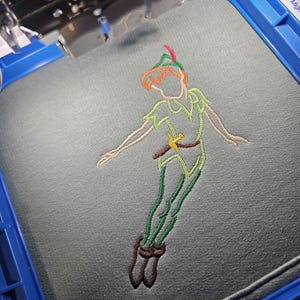 May include: An embroidery machine in action, creating a colourful outline of Peter Pan on grey fabric. The design uses green, orange, and brown thread, showing the character in a flying pose. The embroidery is framed in a blue hoop.