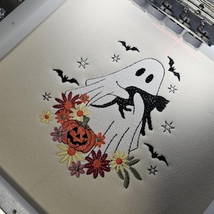 May include: Embroidered design of a white ghost holding a black cat with a pumpkin and flowers. The design is on a white fabric and includes bats and stars.