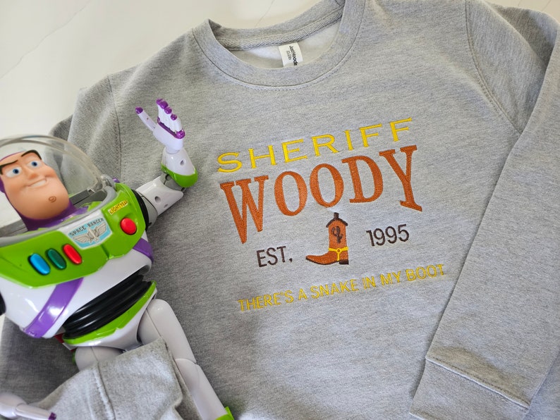 May include: Gray sweatshirt with a yellow, orange, and brown graphic print that says "Sheriff Woody Est. 1995 There's a snake in my boot".