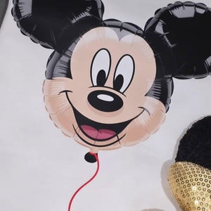 May include: A Mickey Mouse balloon with a smiling face, black ears, and a red string. The balloon is on a white surface, with a black and gold sequined bow in the lower right corner. A party decoration.