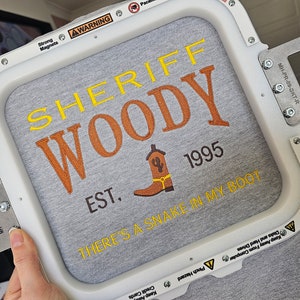 May include: A white embroidery hoop with a gray fabric inside. The fabric has a yellow and orange embroidered design that says "Sheriff Woody Est. 1995 There's a snake in my boot".