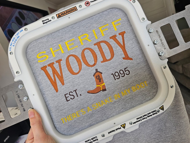 May include: A white embroidery hoop with a gray fabric inside. The fabric has a yellow and orange embroidered design that says "Sheriff Woody Est. 1995 There's a snake in my boot".
