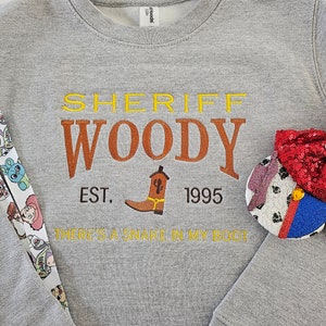 May include: Gray sweatshirt with a yellow, orange, and brown embroidered design that says "Sheriff Woody Est. 1995 There's a snake in my boot".