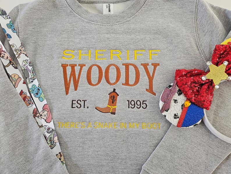 May include: Gray sweatshirt with a yellow, orange, and brown embroidered design that says "Sheriff Woody Est. 1995 There's a snake in my boot".