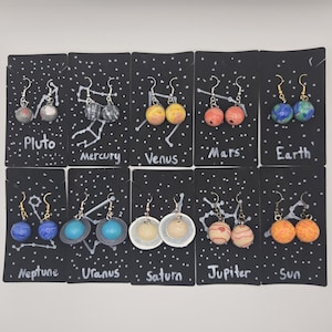 May include: Ten pairs of dangling earrings featuring spherical charms representing the Sun and planets, including Pluto, Mercury, Venus, Mars, Earth, Neptune, Uranus, Saturn, and Jupiter. Each pair is mounted on black cards with white star patterns.