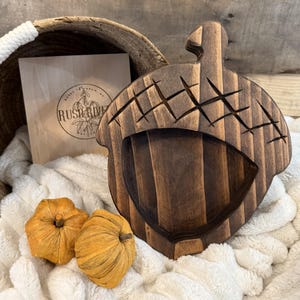 May include: A dark brown wooden acorn-shaped tray with carved details. The tray is next to a wooden box with the text "RUSH RIVER" and two small orange pumpkins. The tray is on a white fluffy blanket.