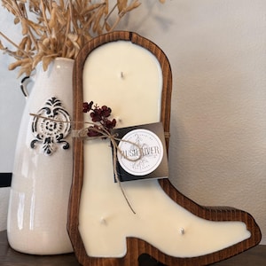 May include: A cowboy boot-shaped candle with a wooden frame. The candle is ivory-colored and has several wicks. A tag with the text "RUSH RIVER" is attached. A decorative vase with dried flowers is in the background.