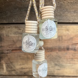 May include: Three glass bottles with wooden tops and a white label that says "Rush River Candles" hanging from a string. The bottles are filled with a clear liquid.
