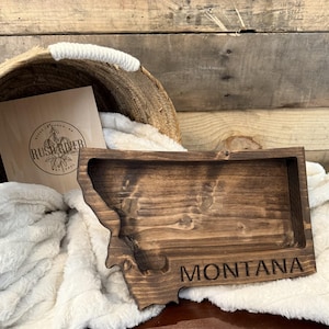 May include: A wooden tray shaped like the state of Montana. The tray is dark brown and has the word "MONTANA" engraved on the bottom.
