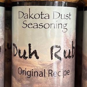May include: A glass jar of Dakota Dust Seasoning, labeled "Duh Rub Original Recipe" with a scenic background image. The jar contains a brown spice blend and has a black lid. The label also states "Net Weight 5.2 oz. (147.4g)".
