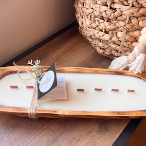 May include: A wooden dough bowl candle with a white wax fill and six wooden wicks. The candle is tied with twine and a small sprig of white flowers. The label on the candle reads "Woodland" with a small floral design.